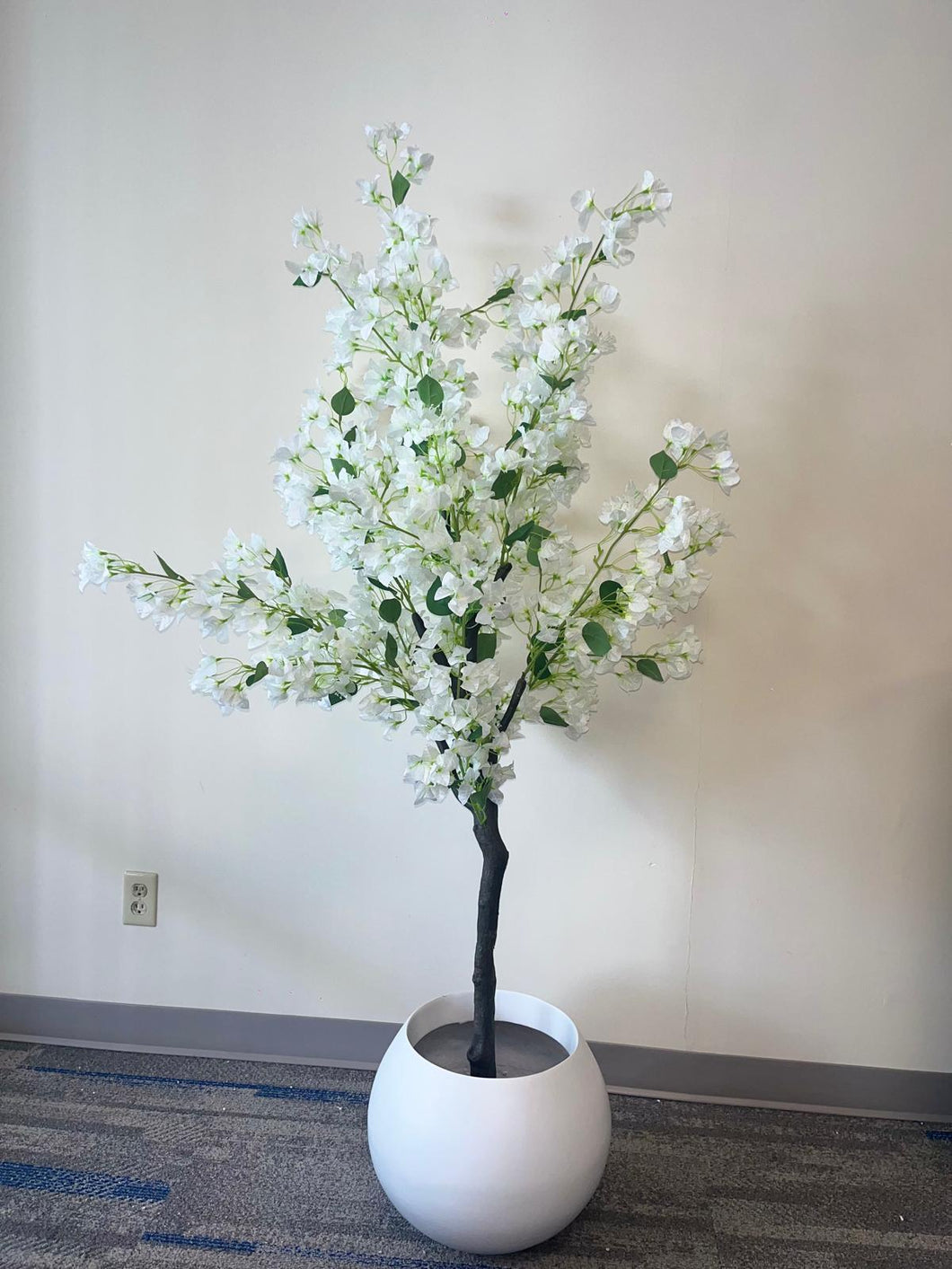 Artificial white flower tree with abundant blossoms in a modern white round planter, placed against a light-colored wall on a carpeted floor