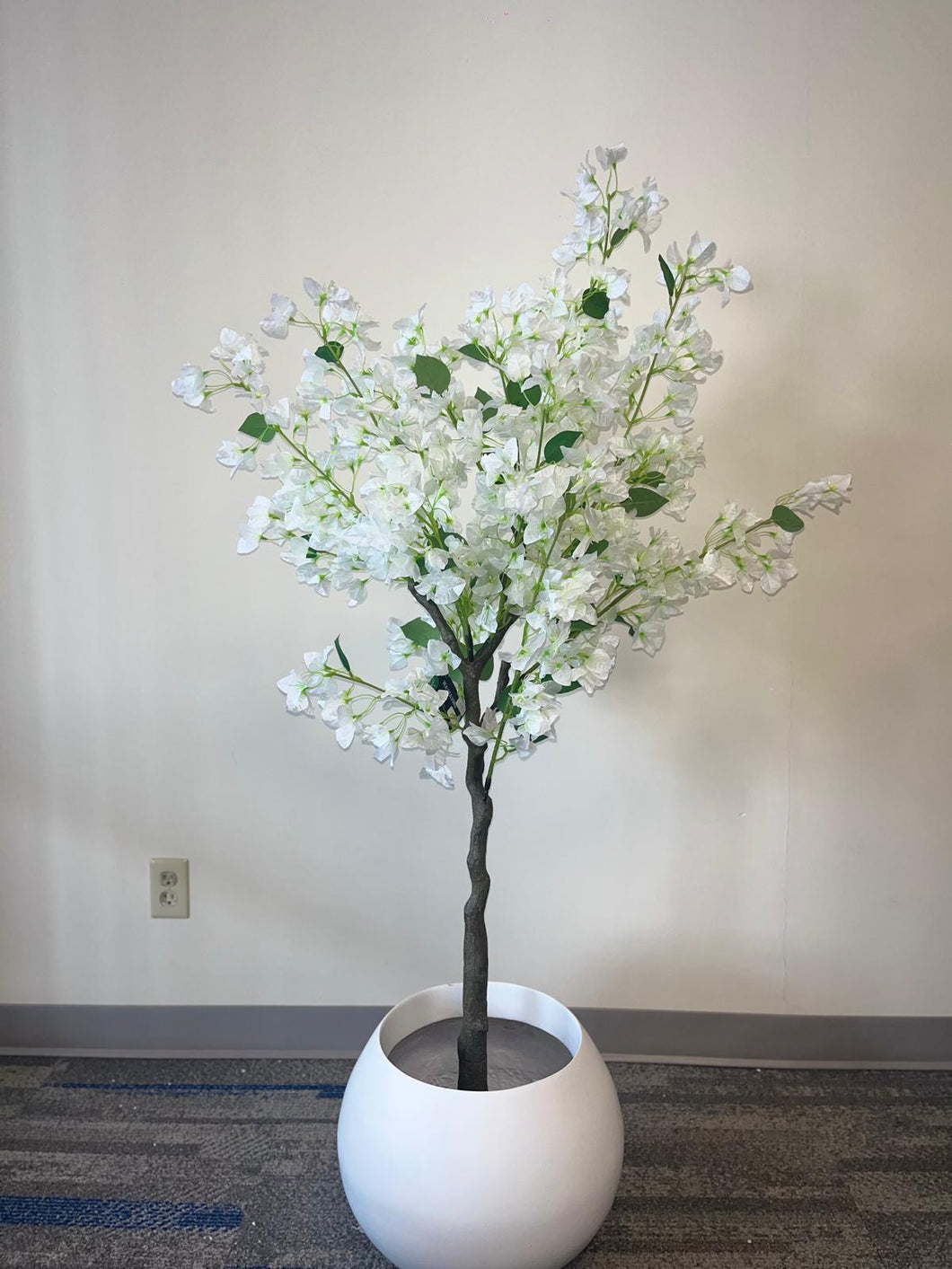 Artificial white flower tree with abundant blossoms in a modern white round planter, placed against a light-colored wall on a carpeted floor