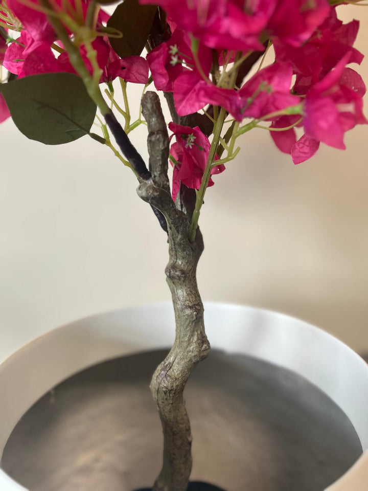 A close-up of an artificial plant trunk emerging from a white pot. The trunk has a textured, realistic bark appearance, with some branches and vibrant pink flowers extending from it. The white pot contrasts with the rich green leaves and vivid pink flowers, creating a striking visual.