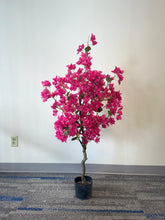 Load image into Gallery viewer, A vibrant artificial plant with bright pink flowers displayed in a compact black pot. The plant stands against a plain wall, with a carpeted floor featuring blue accents. The simple setting allows the vivid pink flowers to be the focal point of the image
