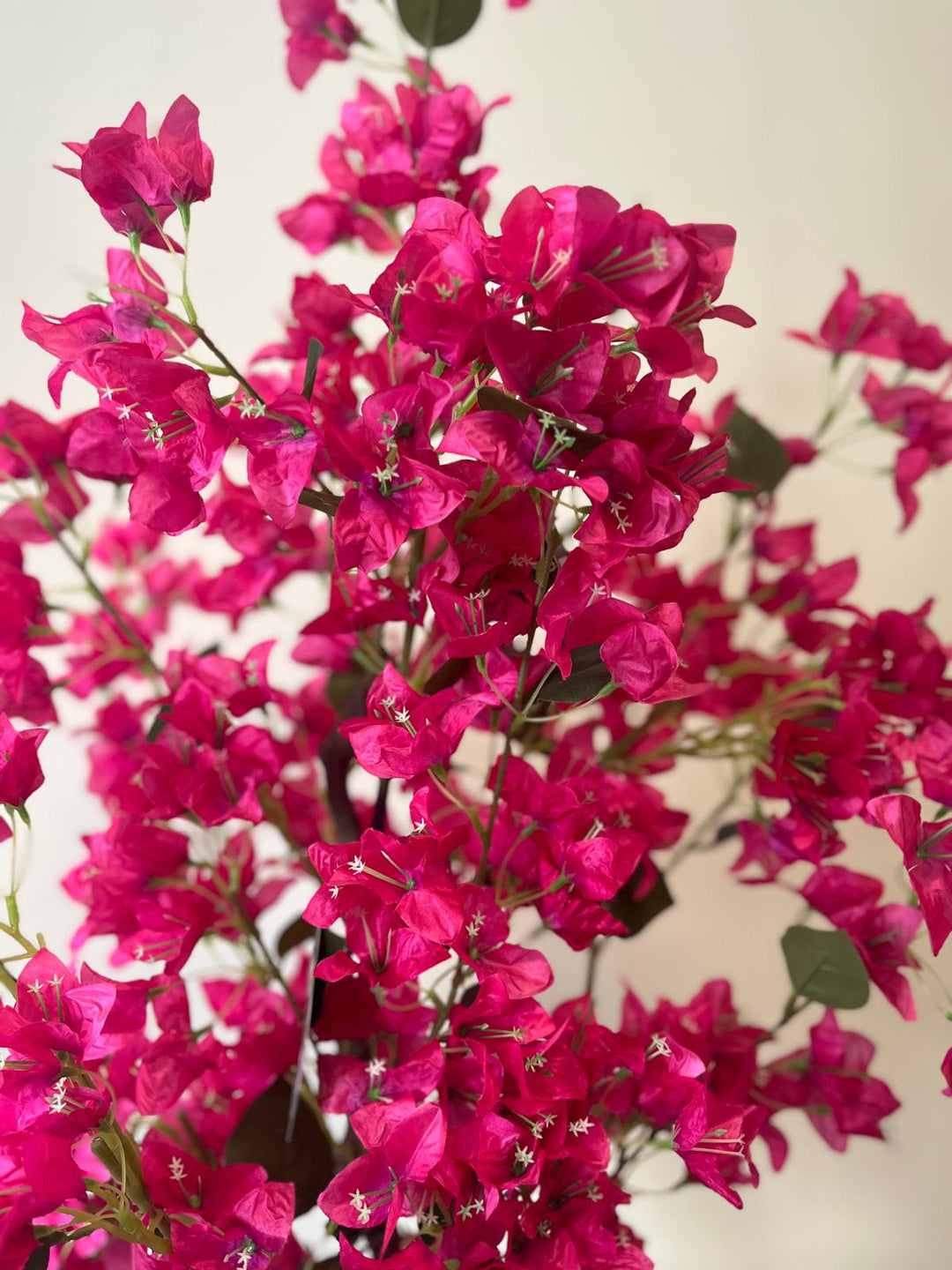 A close-up of a vibrant artificial plant with vivid pink flowers. The petals are detailed, and the small white flower centers are visible. The plant is displayed against a light background, highlighting the rich pink hues and intricate details of the flowers.