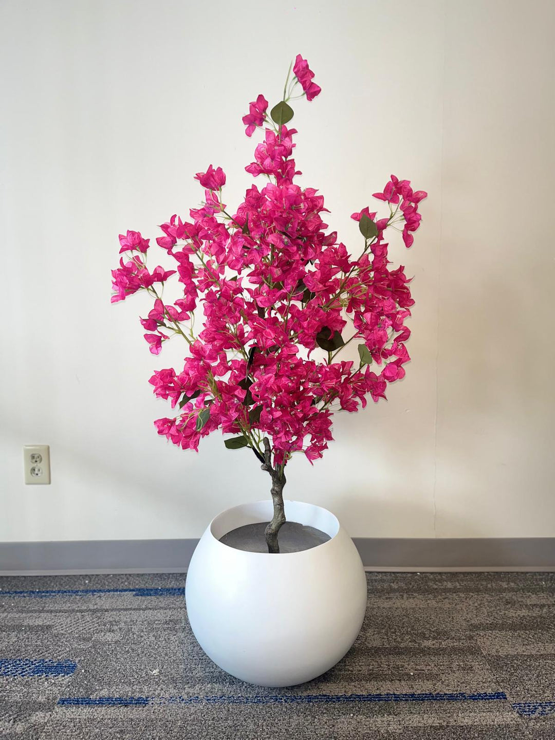 A vibrant artificial plant with bright pink flowers arranged in a full, bushy display, housed in a large, smooth white pot. The plant stands against a plain wall, with a carpeted floor featuring blue accents. The overall setting is simple, allowing the bright pink flowers to stand out prominently