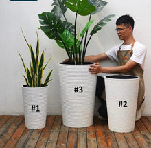 Charger l&#39;image dans la galerie, Person arranging artificial plants, including a snake plant and a monstera in tall, white textured planters of various sizes, on a wooden deck. Common questions: What is the best size planter for large artificial plants? How to choose planters for indoor vs outdoor use? Can these planters be used with real plants?
