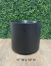 Charger l&#39;image dans la galerie, Matte black cylindrical planter, measuring 11 inches wide by 10 inches high, set on a wooden surface with a green leafy backdrop.
