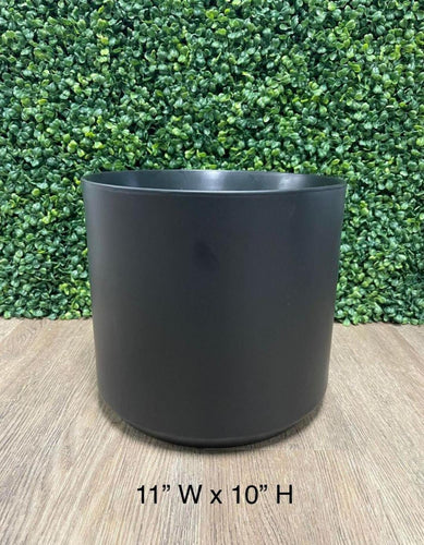 Matte black cylindrical planter, measuring 11 inches wide by 10 inches high, set on a wooden surface with a green leafy backdrop.