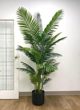 Load image into Gallery viewer, Artificial tall palm tree with lush green fronds placed in a sleek black planter, set against a minimal indoor space with light wood flooring and a white-framed glass door.
