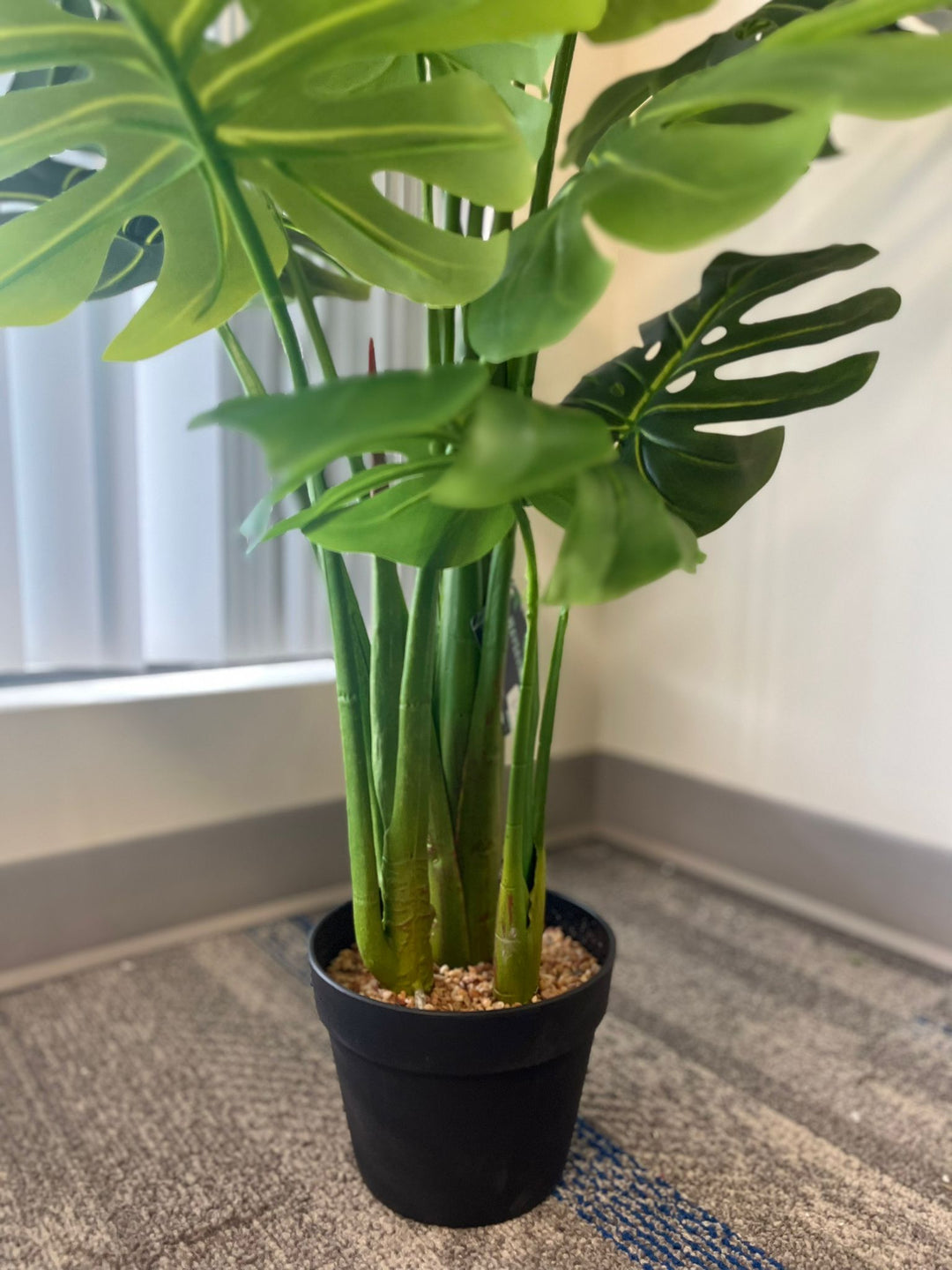 Close-up of the base of an artificial monstera plant showing the green stems emerging from a black pot filled with small pebbles, placed near a window with natural light