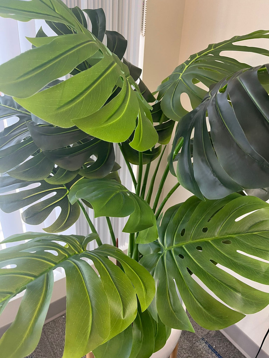 Close-up of artificial monstera leaves showcasing their large, glossy, dark green surface with realistic-looking veins and distinctive splits. The leaves are arranged against a neutral background, emphasizing their lush, tropical appearance.