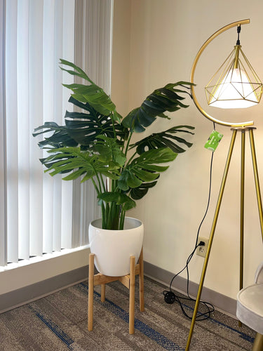 Artificial monstera plant in a white pot with a wooden stand, placed in the corner of a room beside a modern gold floor lamp with a geometric design, near a large window with vertical blinds