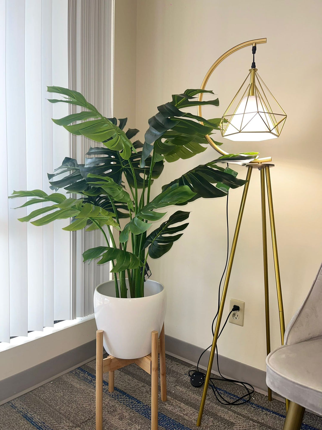 Artificial monstera plant in a white pot with a wooden stand, placed in the corner of a room beside a modern gold floor lamp with a geometric design, near a large window with vertical blinds.