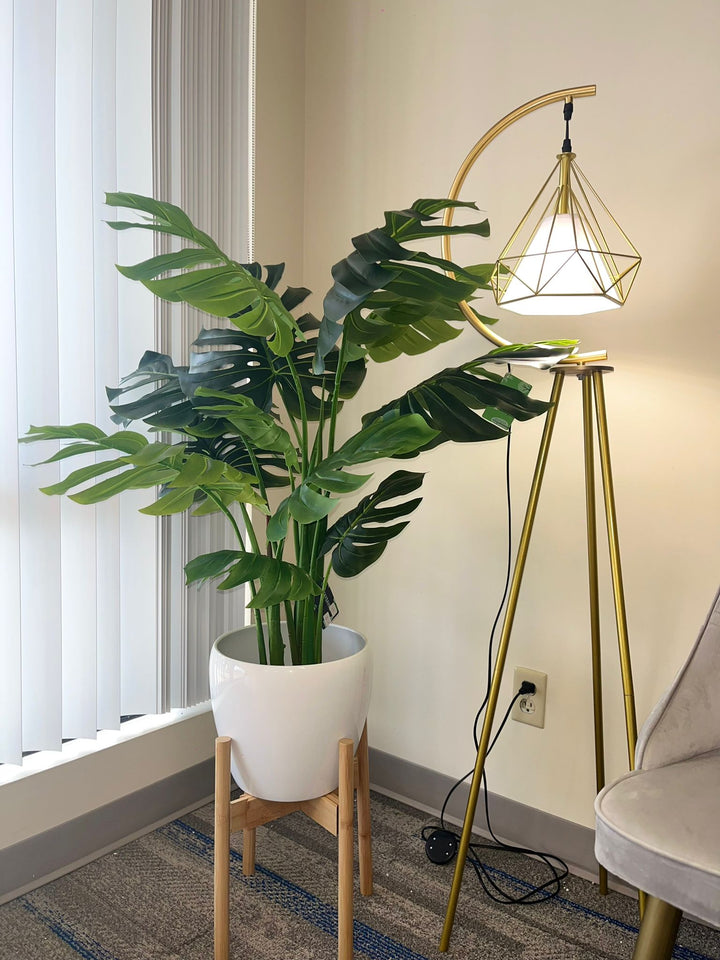 Artificial monstera plant in a white pot with a wooden stand, placed in the corner of a room beside a modern gold floor lamp with a geometric design, near a large window with vertical blinds.