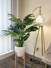 Charger l&#39;image dans la galerie, Artificial monstera plant in a white pot with a wooden stand, placed in the corner of a room beside a modern gold floor lamp with a geometric design, near a large window with vertical blinds.
