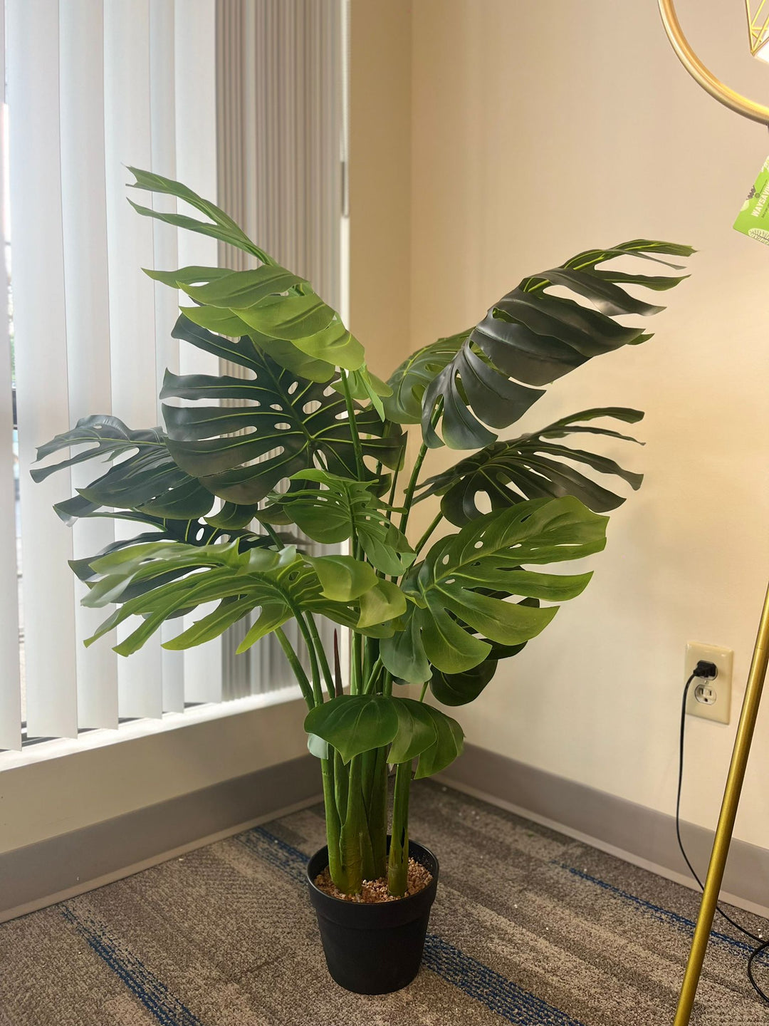Artificial monstera plant in a white pot with a wooden stand, placed in the corner of a room beside a modern gold floor lamp with a geometric design, near a large window with vertical blinds