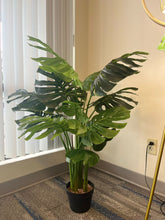 Charger l&#39;image dans la galerie, Artificial monstera plant in a white pot with a wooden stand, placed in the corner of a room beside a modern gold floor lamp with a geometric design, near a large window with vertical blinds
