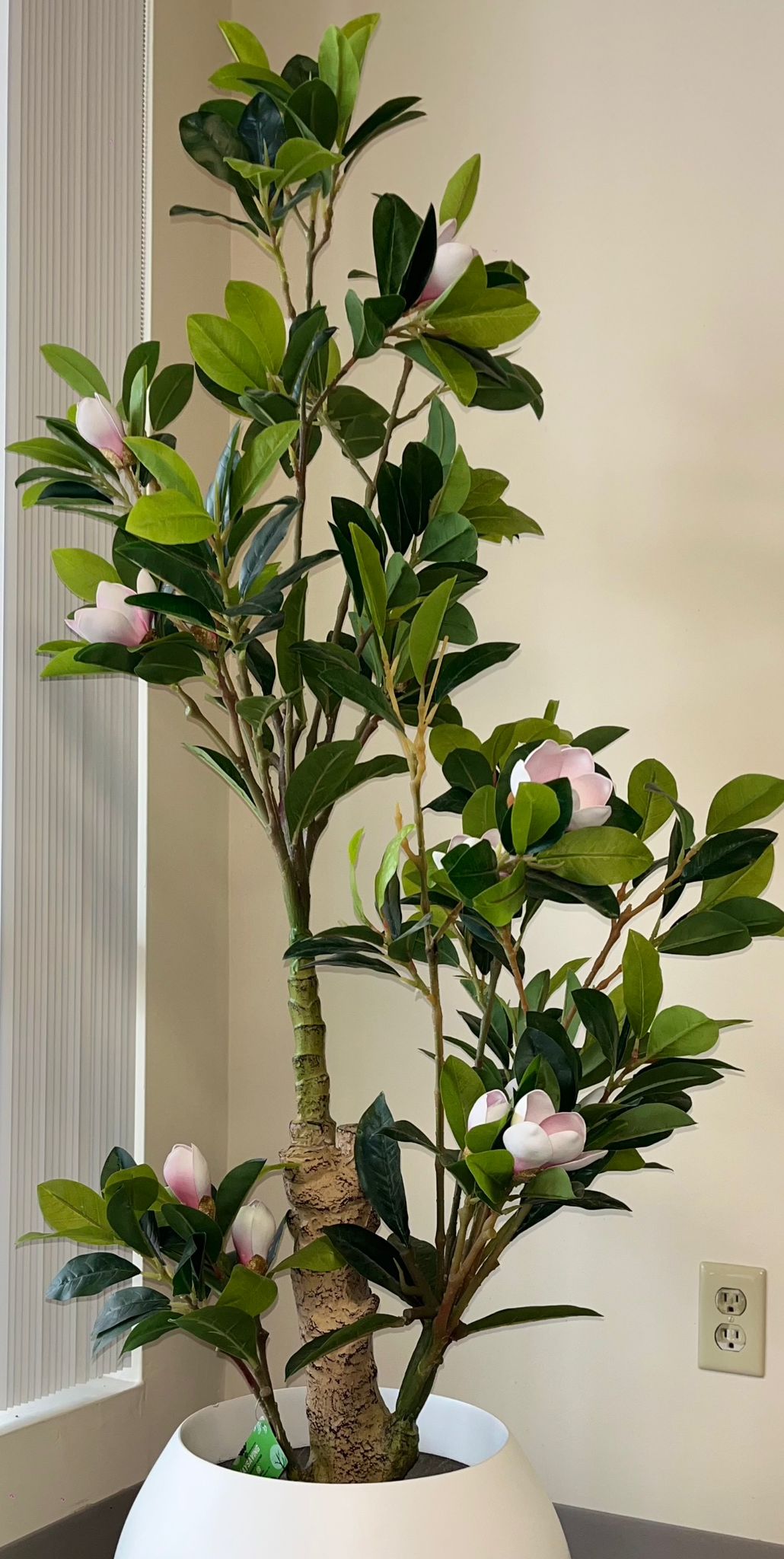 Artificial indoor tree with green leaves and pink flowers in a white round pot, placed in a modern interior setting with neutral tones.