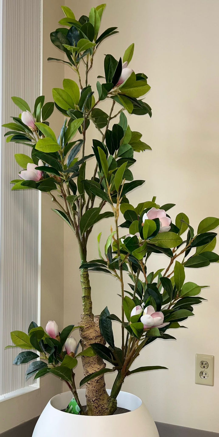 Artificial indoor tree with green leaves and pink flowers in a white round pot, placed in a modern interior setting with neutral tones.