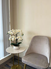 Load image into Gallery viewer, Minimalist corner of a modern room featuring a light gray upholstered chair paired with a small, round side table with a white marble top and gold base. The table holds a white pot with elegant white orchids, enhancing the serene and sophisticated decor. Light-colored walls and vertical blinds in the background contribute to the clean and peaceful ambiance, ideal for relaxation. Perfect interior design inspiration for a cozy reading nook or minimalist home office setup.
