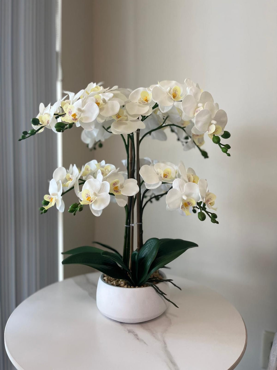 This image shows an elegant arrangement of artificial white orchids placed on a round, white marble-top table. The orchids are beautifully arranged in a white, smooth, rounded pot, with green leaves and a few visible buds, making the flowers appear lush and lifelike. The flowers have delicate white petals with soft yellow and pink centers, contributing to a serene and sophisticated aesthetic. The background is minimal, allowing the focus to remain on the orchid arrangement.