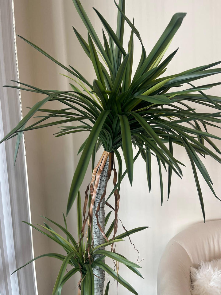 Close-up view of a tall artificial dracaena plant with realistic green leaves and brown decorative ribbon accents on the trunk, providing a detailed look at its lifelike design for home or office decor