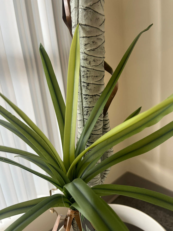 Close-up shot of the base of an artificial dracaena plant, featuring realistic green leaves and a textured faux trunk, showcasing the intricate detail for indoor home or office decor.