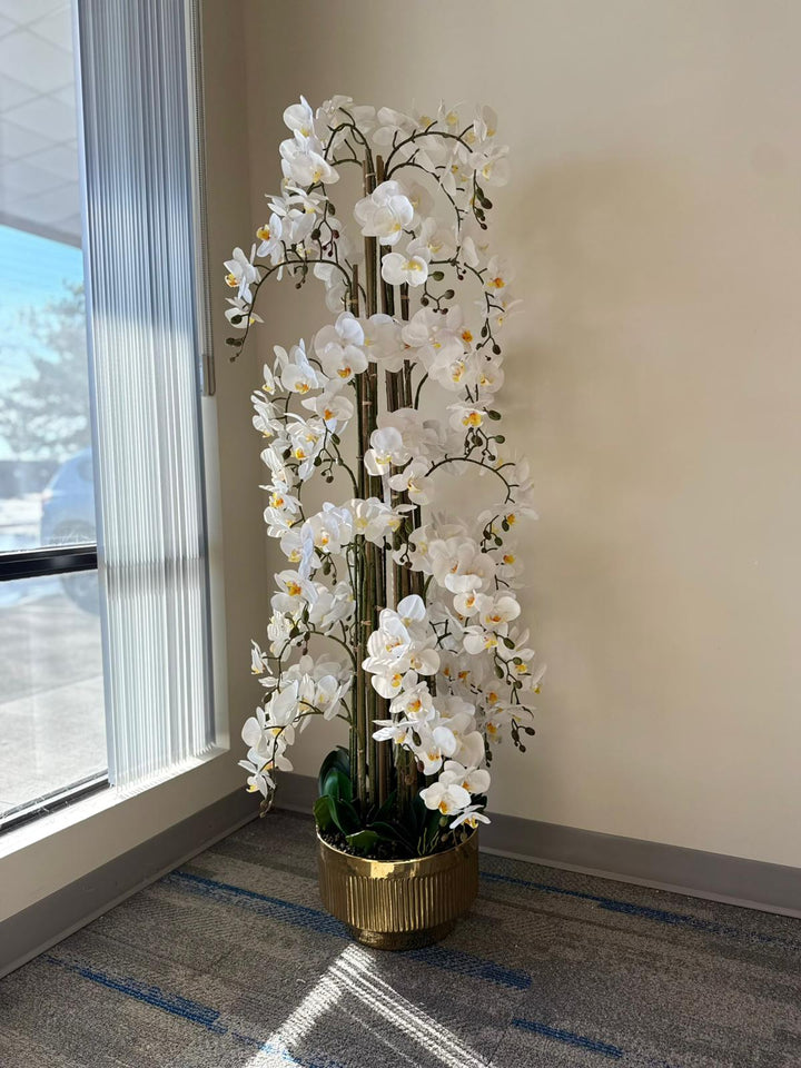 Tall artificial white orchid plant in a gold pot on a carpeted floor near a window.
