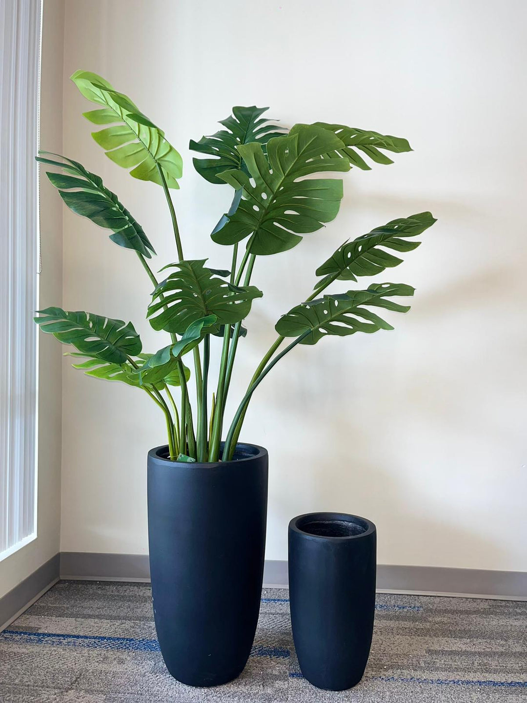 Set of two black matte cylindrical planters. The larger planter showcases a lush, artificial monstera plant with vibrant green leaves, perfect for enhancing any indoor space. The planters' minimalist design adds a modern touch to any decor. Ideal for home or office settings.