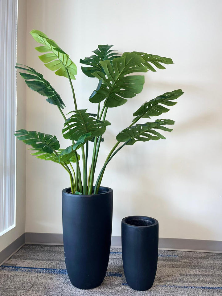 Set of two black matte cylindrical planters. The larger planter showcases a lush, artificial monstera plant with vibrant green leaves, perfect for enhancing any indoor space. The planters' minimalist design adds a modern touch to any decor. Ideal for home or office settings.