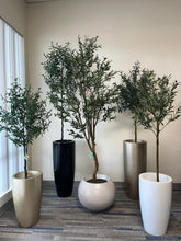 Charger l&#39;image dans la galerie, A group of artificial olive trees placed in modern planters. The planters are tall and sleek in black, white, gold, and silver, with one round, white pot on the floor. The trees feature realistic green leaves and are positioned against a beige wall, near a window with vertical blinds, creating a clean and elegant indoor display.
