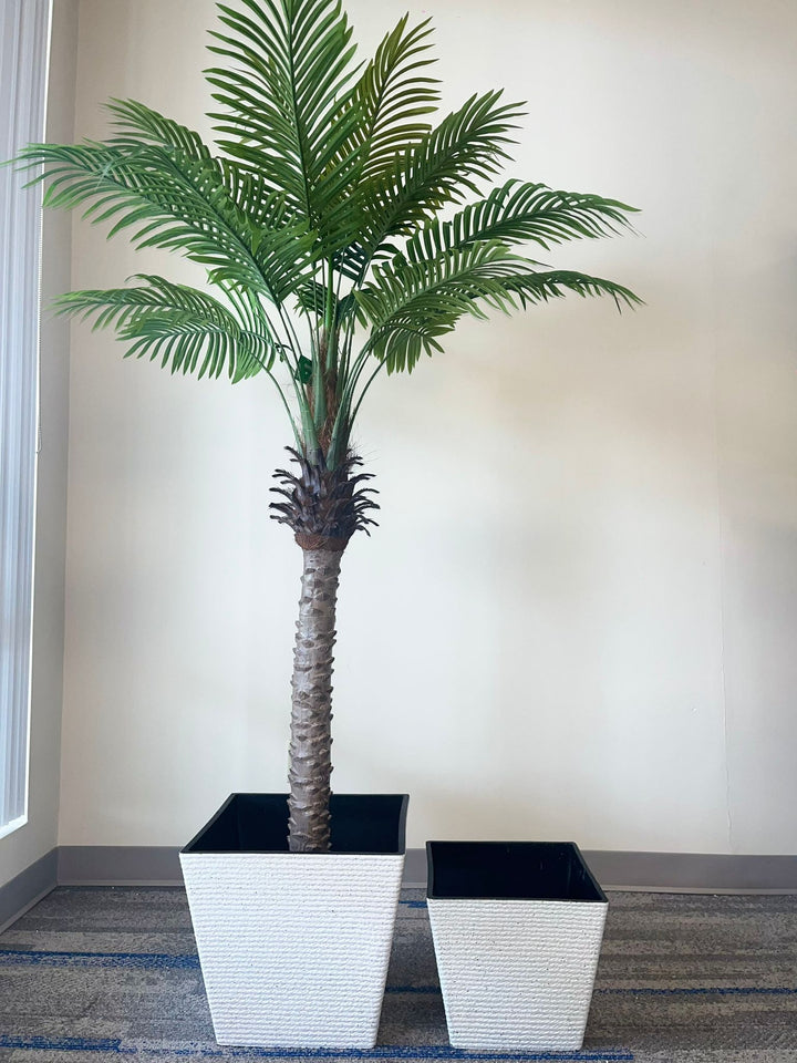 A tall artificial palm tree placed inside a large white square textured planter with a black interior. A smaller, empty planter of the same design is positioned next to it on the right. Both planters rest on a carpeted floor against a plain wall, creating a modern indoor setting.