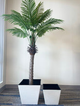 Load image into Gallery viewer, A tall artificial palm tree placed inside a large white square textured planter with a black interior. A smaller, empty planter of the same design is positioned next to it on the right. Both planters rest on a carpeted floor against a plain wall, creating a modern indoor setting.
