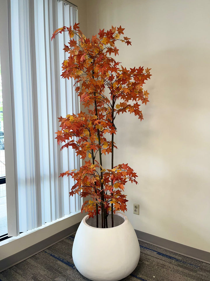 A tall artificial tree with vibrant orange and red autumn leaves, planted in a round white planter, placed in a corner by a large window with vertical blinds.