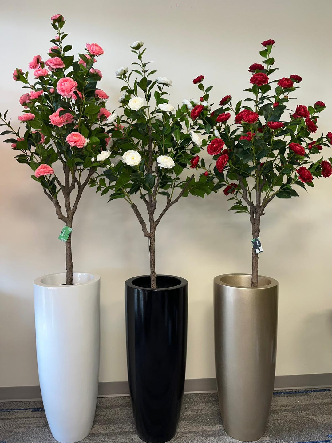 Three artificial flowering trees are displayed in sleek, tall planters in white, black, and gold. The trees feature vibrant pink, white, and red blossoms, creating a striking contrast against the green leaves. The arrangement adds a pop of color to the minimalist indoor space with a neutral beige wall as the background.