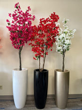 Charger l&#39;image dans la galerie, Three artificial flowering trees are displayed in sleek, tall planters in white, black, and gold. The trees feature vibrant pink, white, and red blossoms, creating a striking contrast against the green leaves. The arrangement adds a pop of color to the minimalist indoor space with a neutral beige wall as the background.
