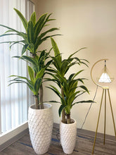 Charger l&#39;image dans la galerie, Two artificial plants with broad green leaves in tall, white textured planters placed beside a window with vertical blinds. How can large artificial plants be used to fill empty spaces in modern decor?
