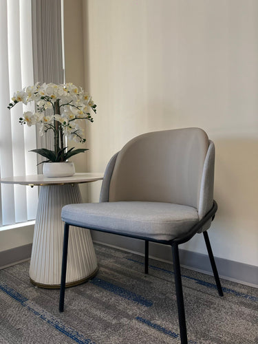 Modern chair with light gray cushioned seat and back, paired with a small round side table. The table features a white pleated base and a marble top, adorned with an artificial white orchid flower arrangement in a minimalist space.