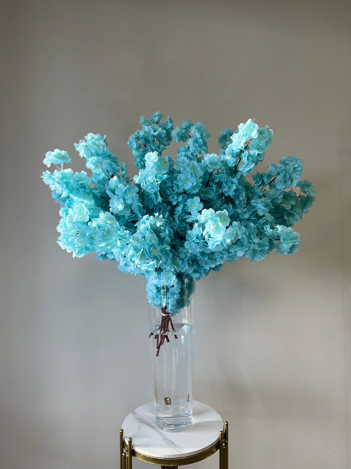 Elegant artificial turquoise flowers beautifully arranged in a tall clear glass vase filled with water, set on a marble pedestal. How can these vibrant blooms enhance your wedding or event décor?