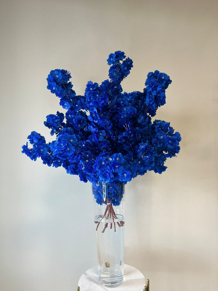 Vibrant blue artificial flowers elegantly arranged in a tall, clear glass vase filled with water, showcased on a chic marble and gold pedestal. Ideal for weddings, events, and upscale décor. How can these striking blue blooms transform your event?