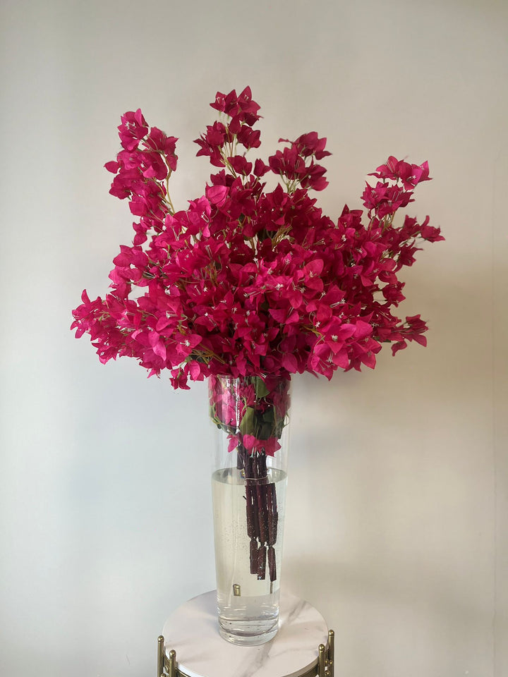 Vibrant artificial magenta bougainvillea flowers arranged in a tall clear glass vase filled with water, elegantly placed on a marble pedestal. What makes this arrangement ideal for modern event décor?