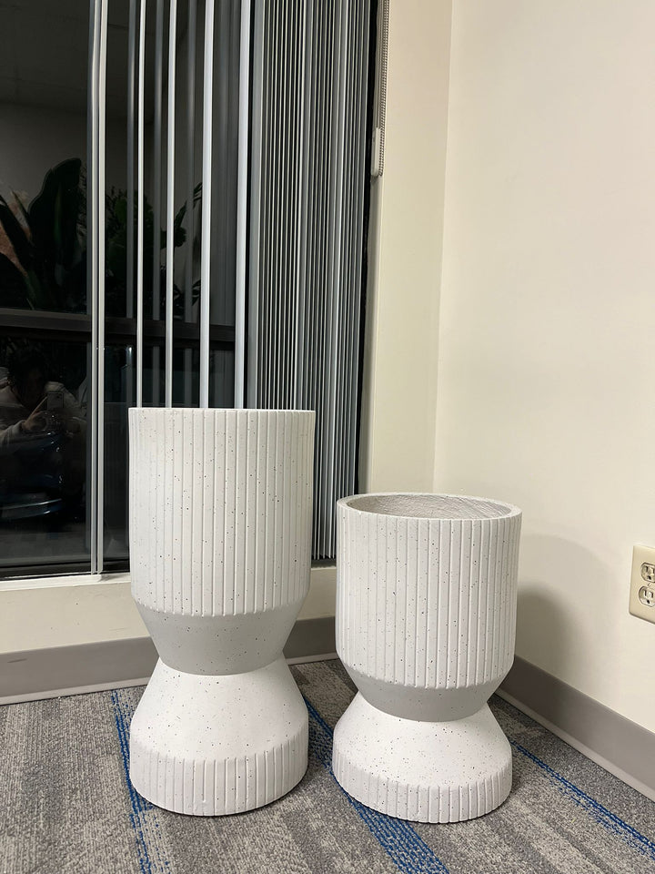 Set of two white ribbed cylindrical planters with a modern textured design, displayed empty indoors near a window on a carpeted floor. Are these planters suitable for heavy plants?