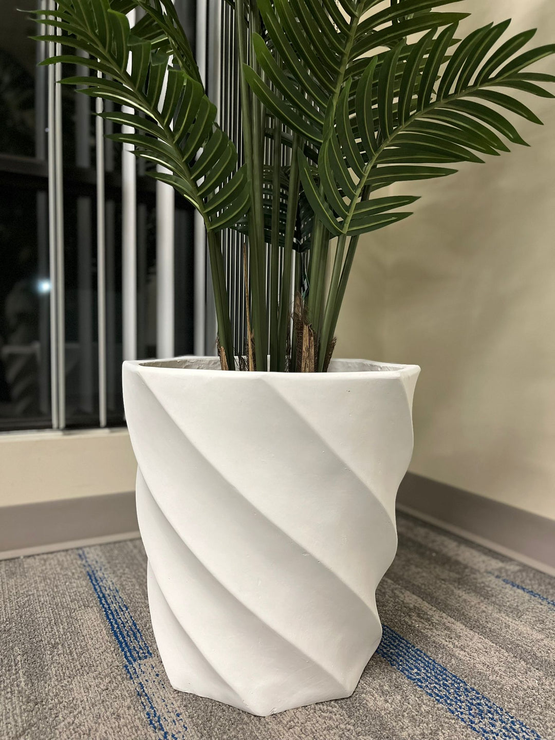 modern white spiral-patterned planter containing a lush green plant with long, elegant fronds, placed on a carpeted floor near a window with vertical blinds.