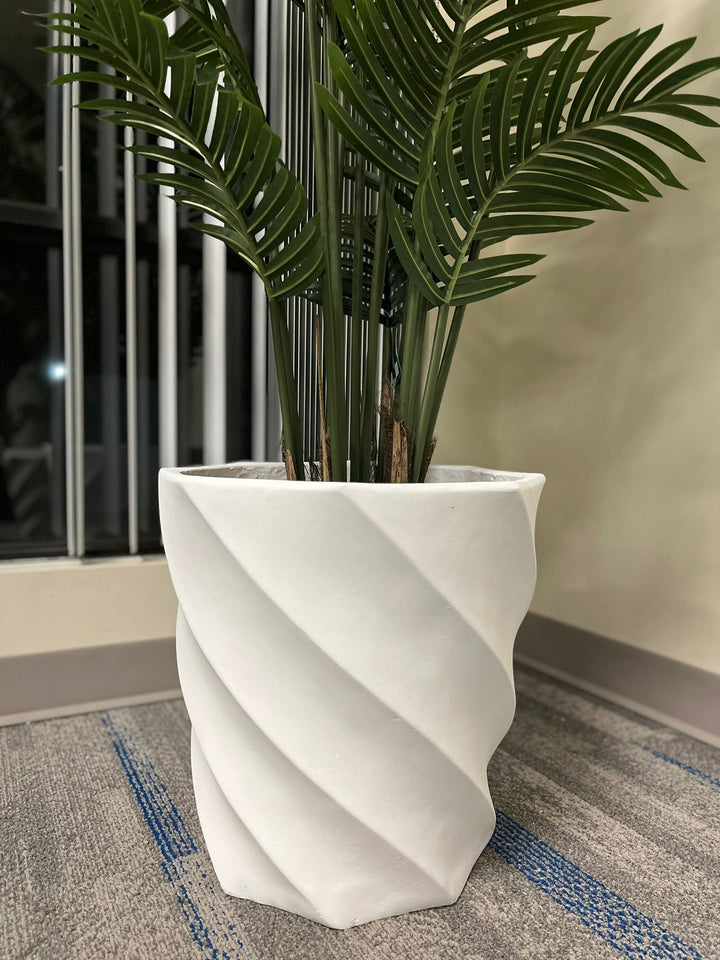 modern white spiral-patterned planter containing a lush green plant with long, elegant fronds, placed on a carpeted floor near a window with vertical blinds.