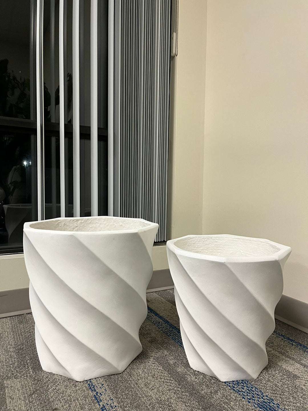 white, contemporary spiral-designed planters of varying sizes, placed side by side indoors on a carpeted floor near vertical blinds, showcasing a sleek and modern aesthetic.