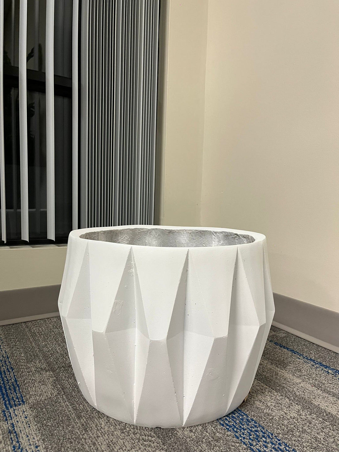 White planter with a faceted geometric design, displayed empty indoors near a window on a carpeted floor. Does this planter include a drainage hole for excess water?
