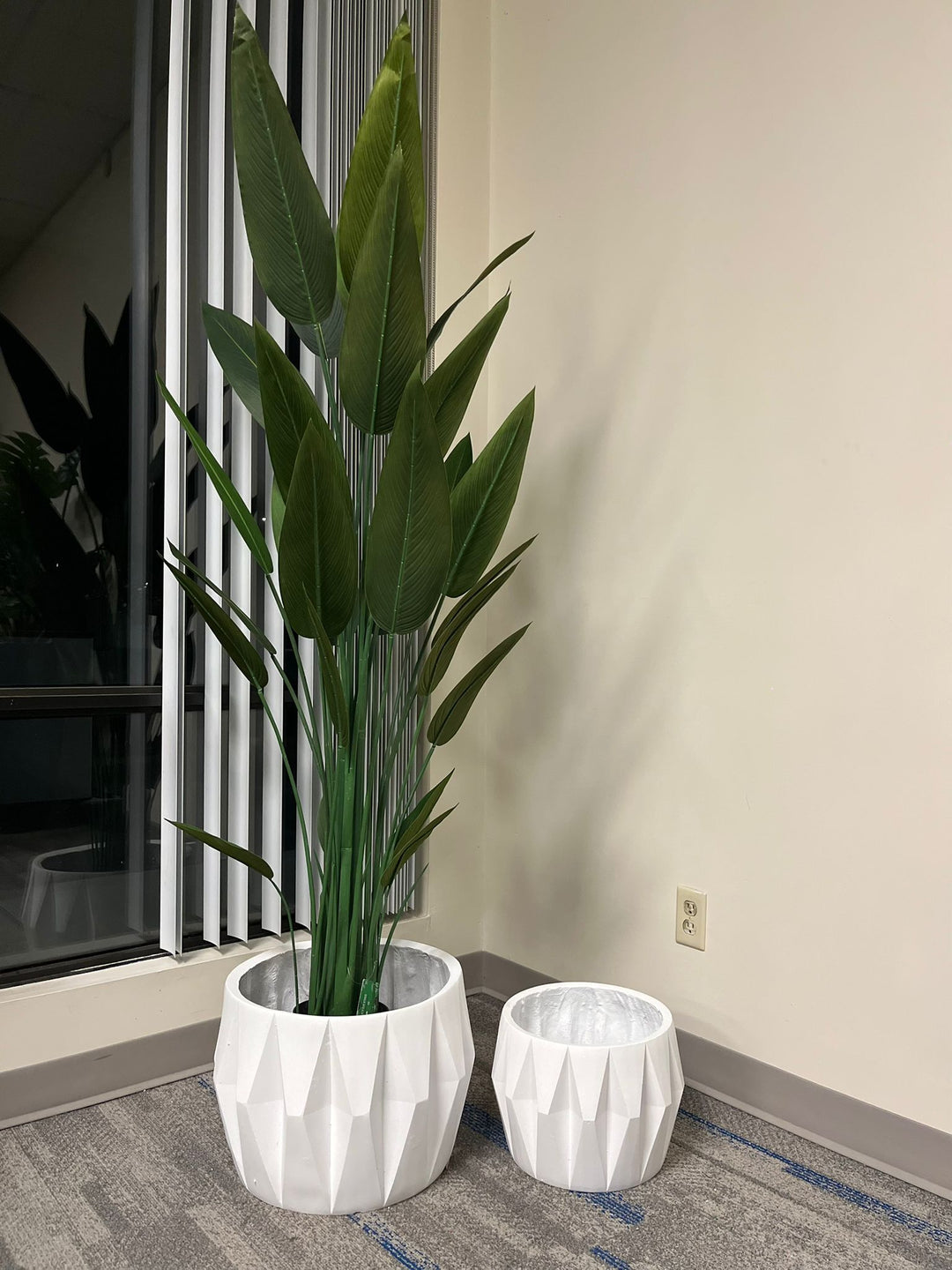 Set of two white faceted geometric planters, one holding a tall artificial plant with lush green leaves, placed indoors near a window on a carpeted floor. Are these planters made of durable materials for long-term use?