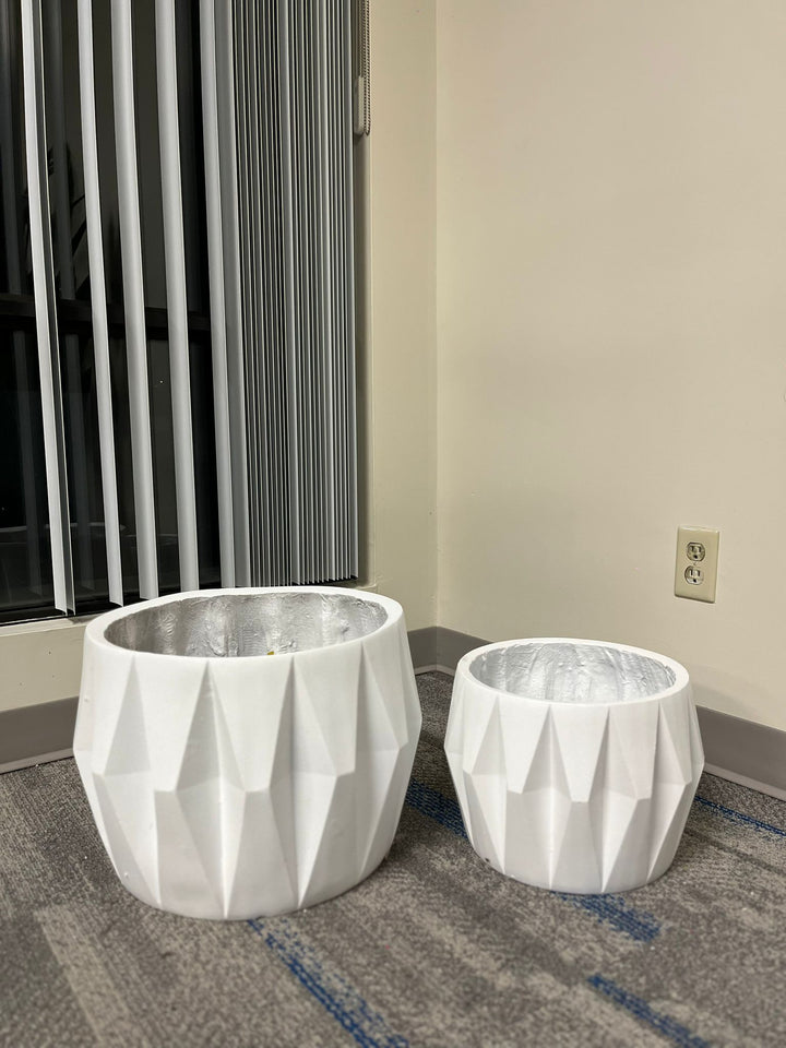 Set of two white planters with a faceted geometric design, displayed empty indoors near a window on a carpeted floor. Are these planters available in different sizes?