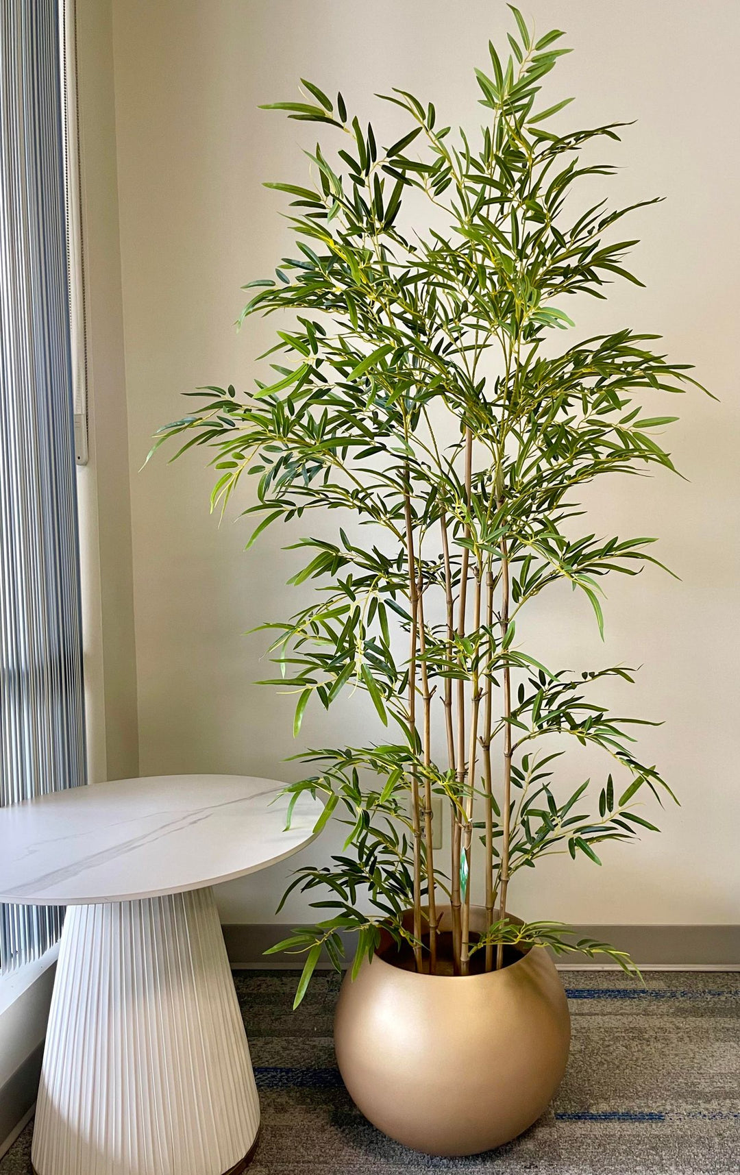 Tall artificial bamboo plant in a round gold pot, positioned next to a modern white side table.