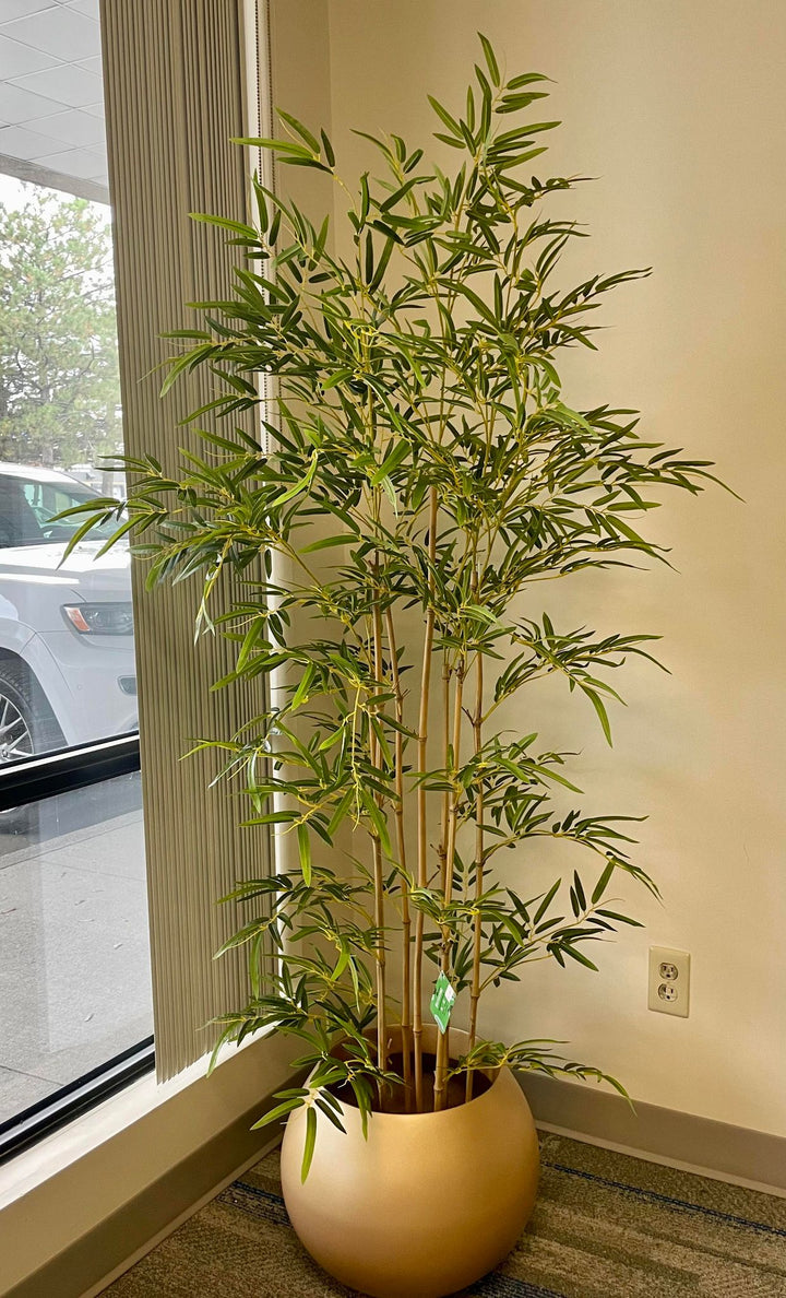 Artificial bamboo plant in a gold pot, placed near a window in a well-lit room adding a realistic greenery to the setting.