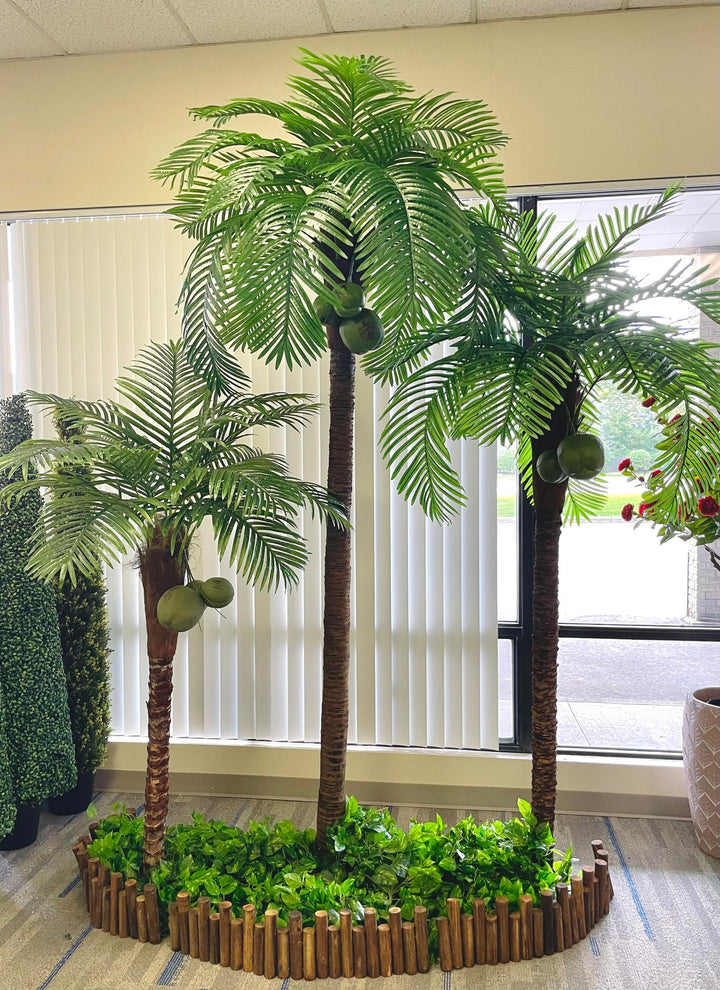 Artificial palm trees with coconuts set up indoors, surrounded by lush greenery and enclosed in a wooden planter, creating a vibrant tropical display.
