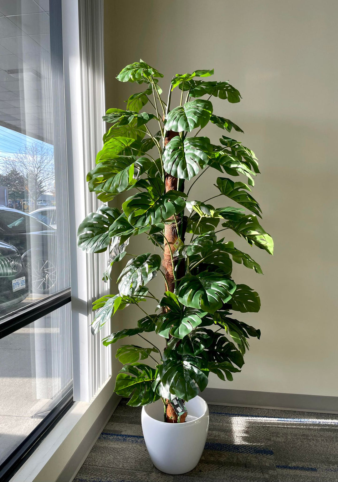 Artificial monstera plant in a white pot placed beside a modern window in a stylish living room. The lush green foliage adds a vibrant touch to the neutral-colored space.