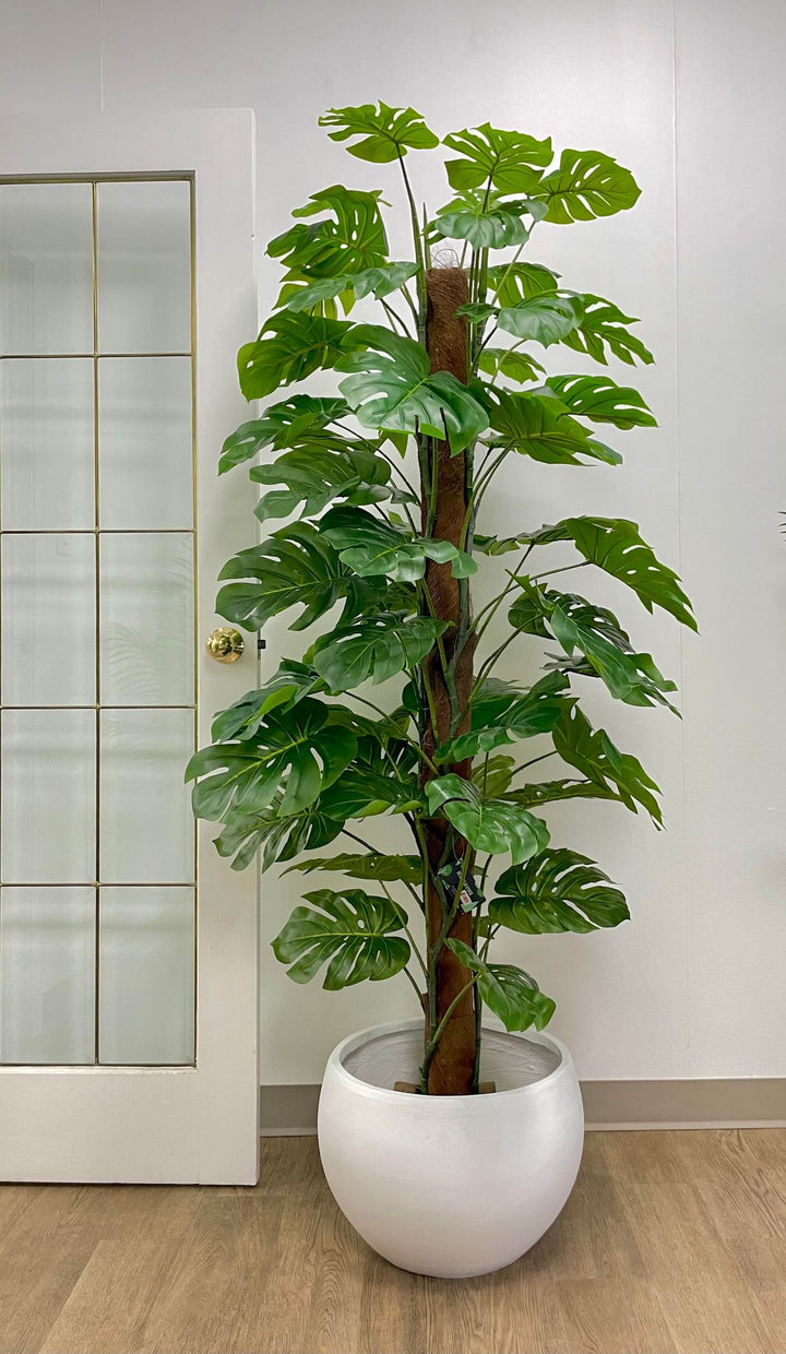 Tall artificial monstera plant placed in a sleek white planter near a window. The lush green leaves with natural-looking veins add a tropical vibe to the indoor setting.